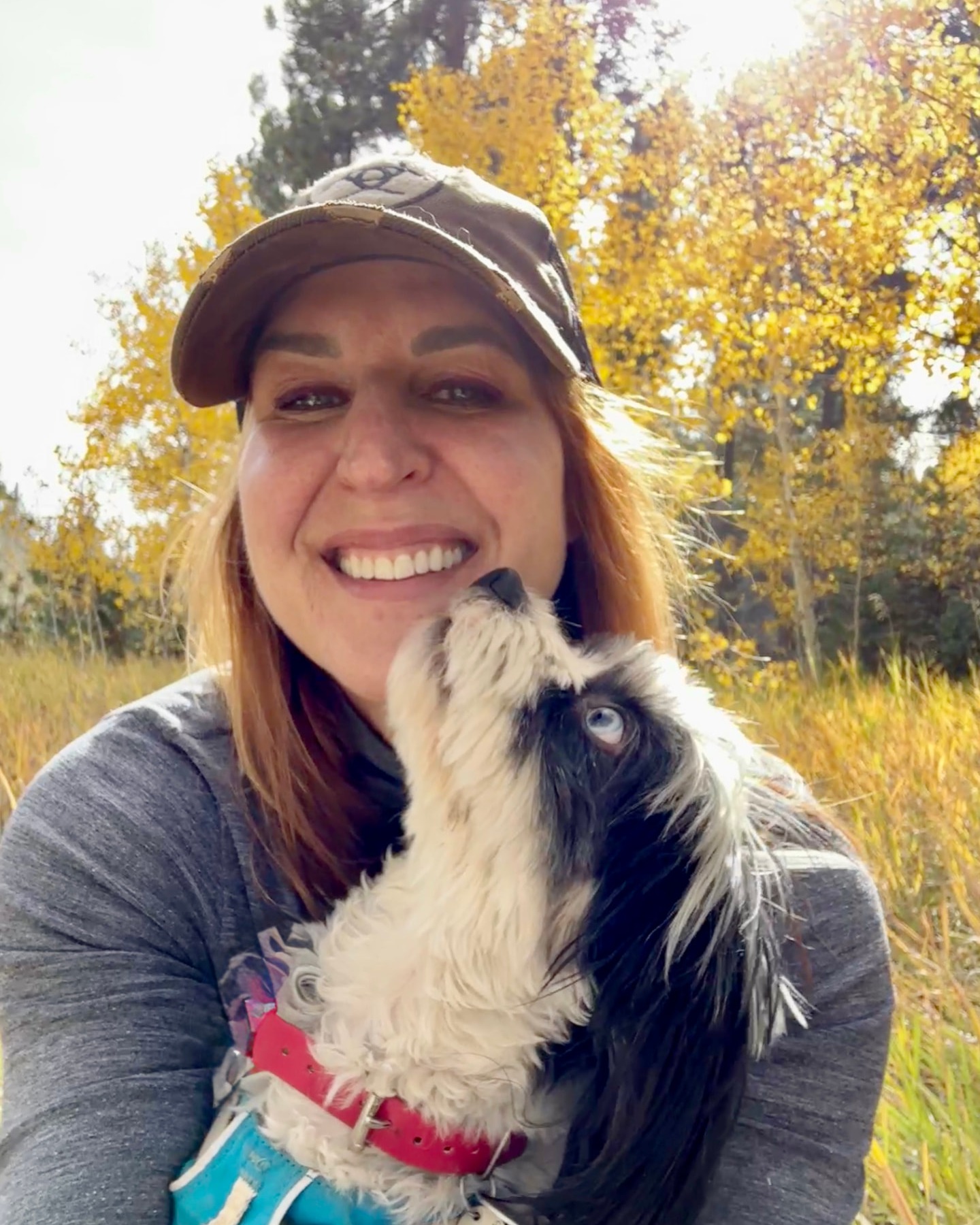 Portrait of Marylou smiling outdoors with a small black and white dog in her arms and golden aspen trees in the background